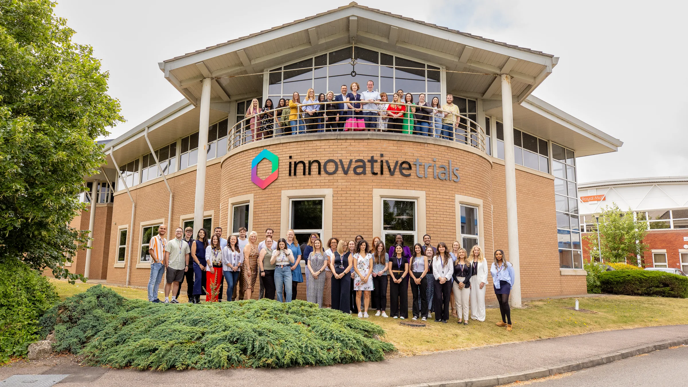 The Innovative Trials team outside Innovative House, some on the balcony under the company sign and the rest on the lawn below