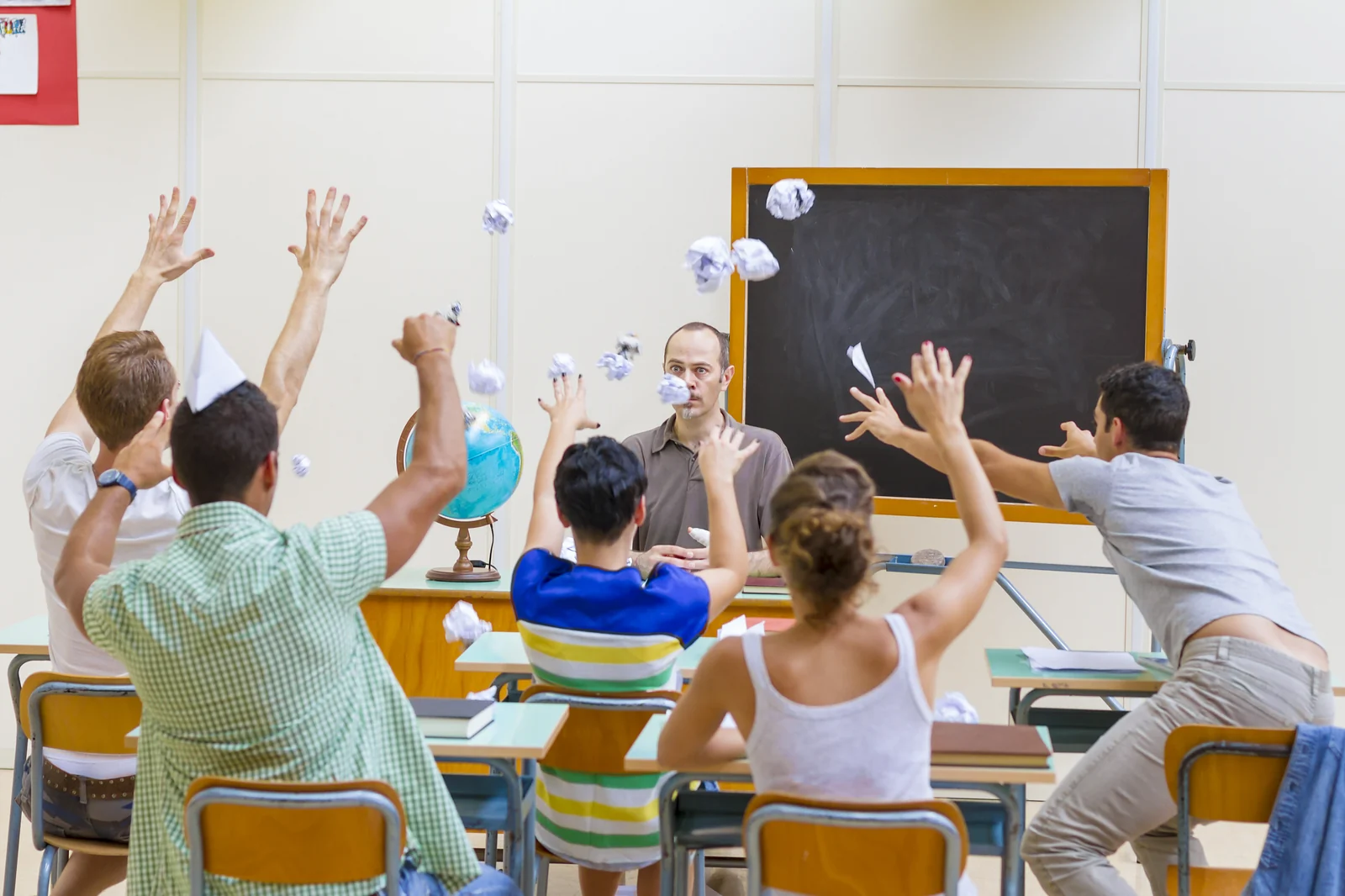 A classroom of students throwing paper balls in the air in front of their teacher at the blackboard