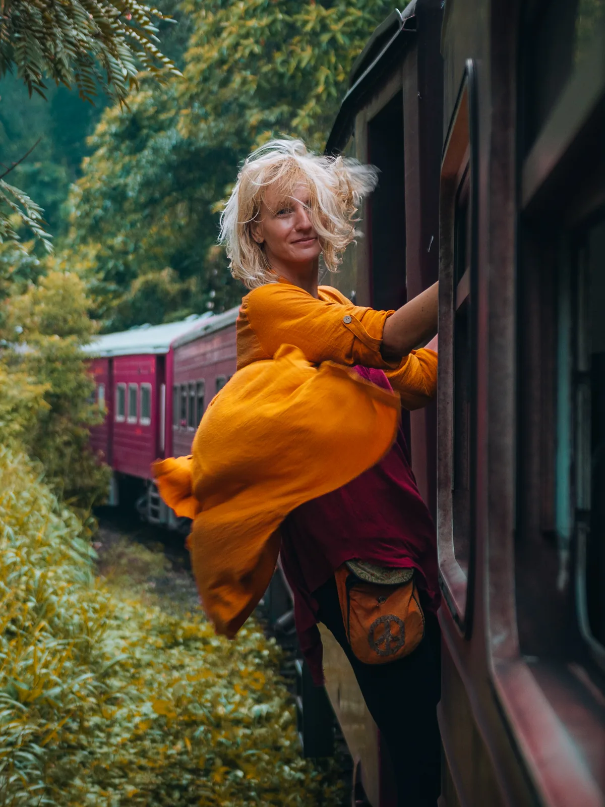 A woman in an orange scarf leaning out of a moving train, smiling back at the camera