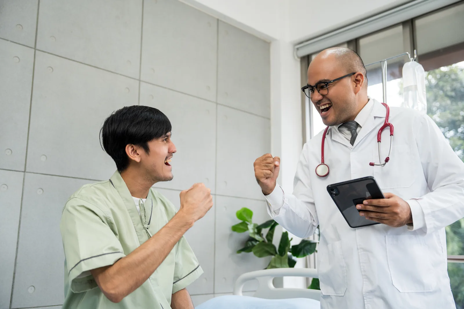 A doctor with a tablet and a patient sitting on a hospital bed, both celebrating with raised fists