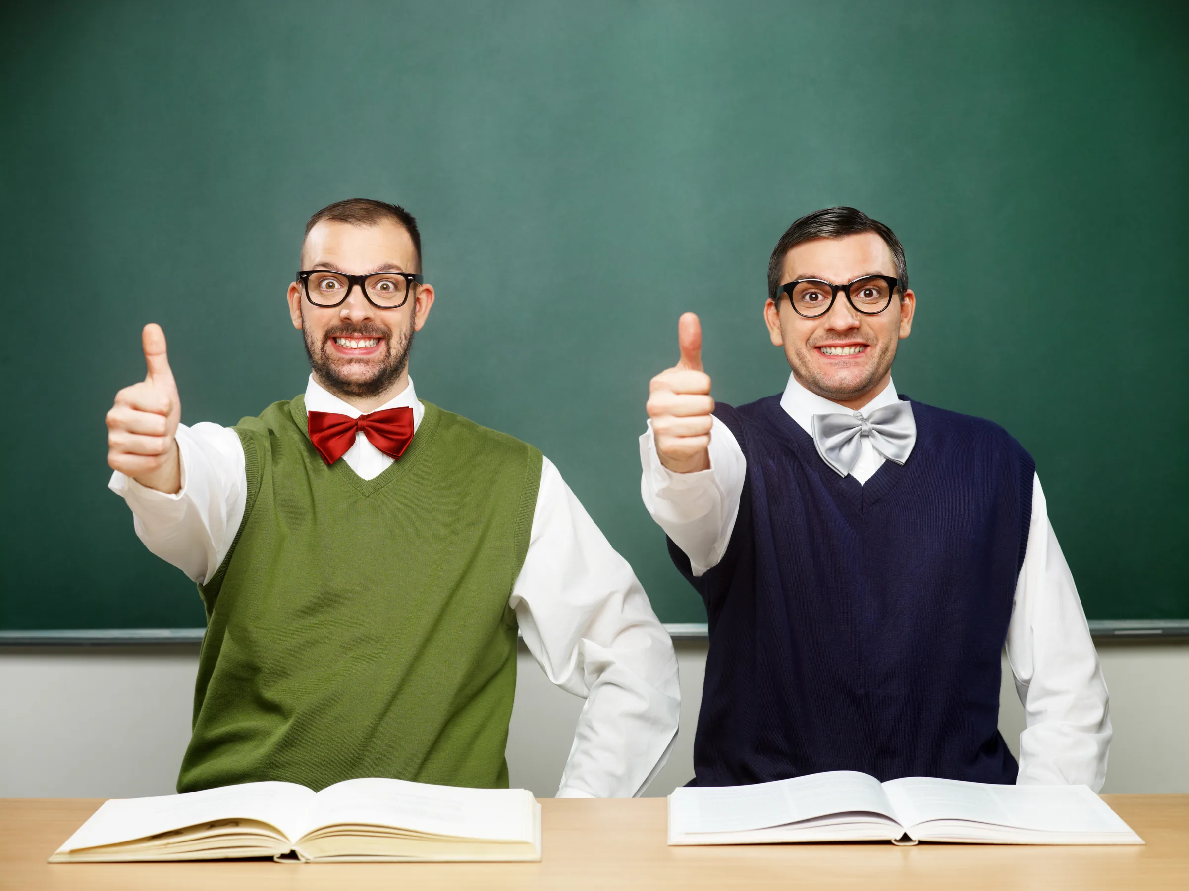 Two men in glasses and bow ties at a classroom desk, giving a thumbs up in front of a blackboard