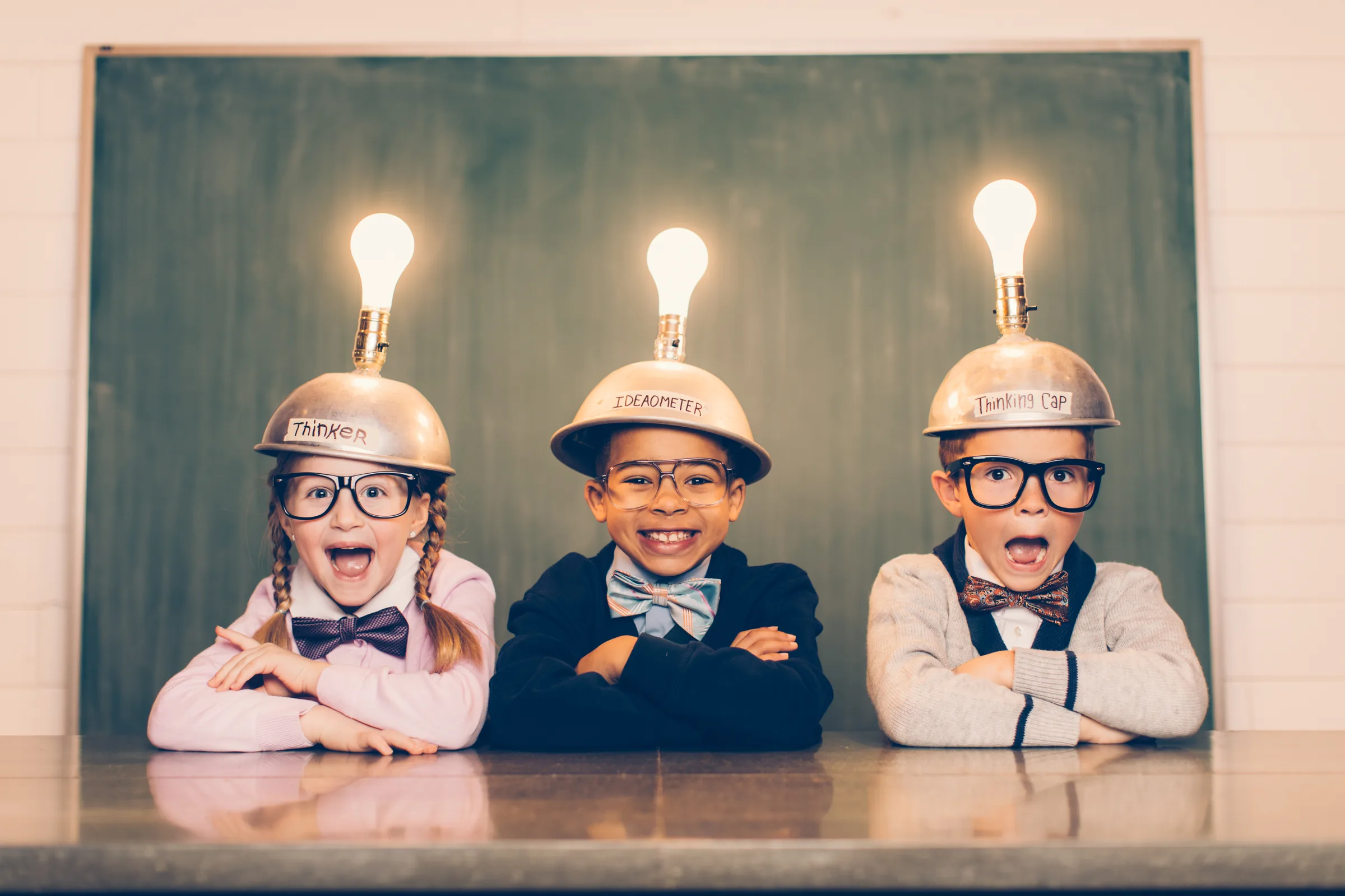 Three children wearing lit-up lightbulb helmets, grinning at their desks