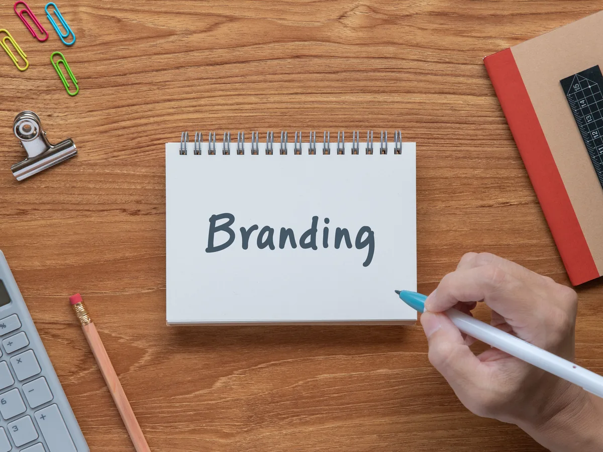 A hand writing on a spiral notepad that reads Branding, on a wooden desk