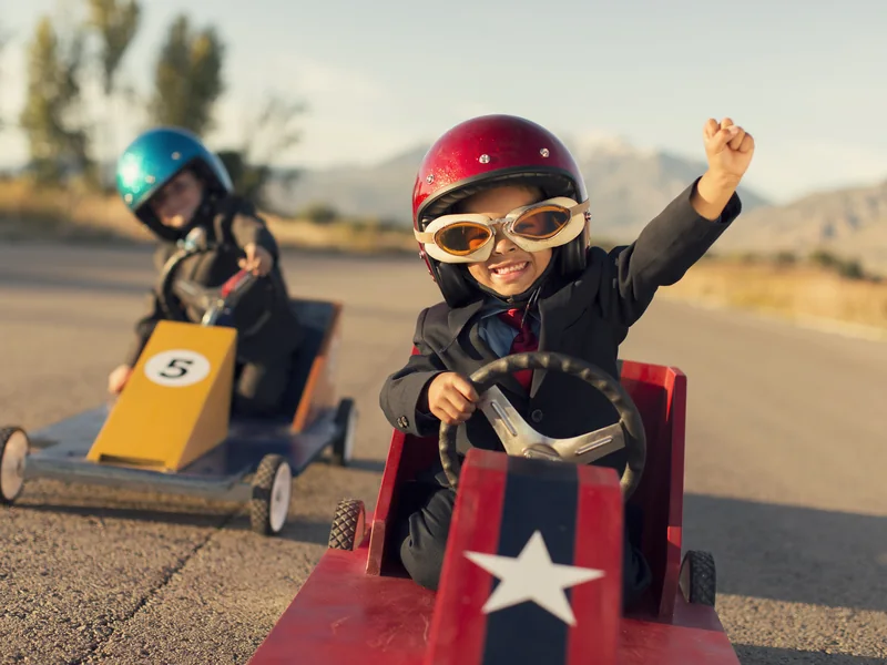Two children in suits and helmets racing go-karts, one punching the air