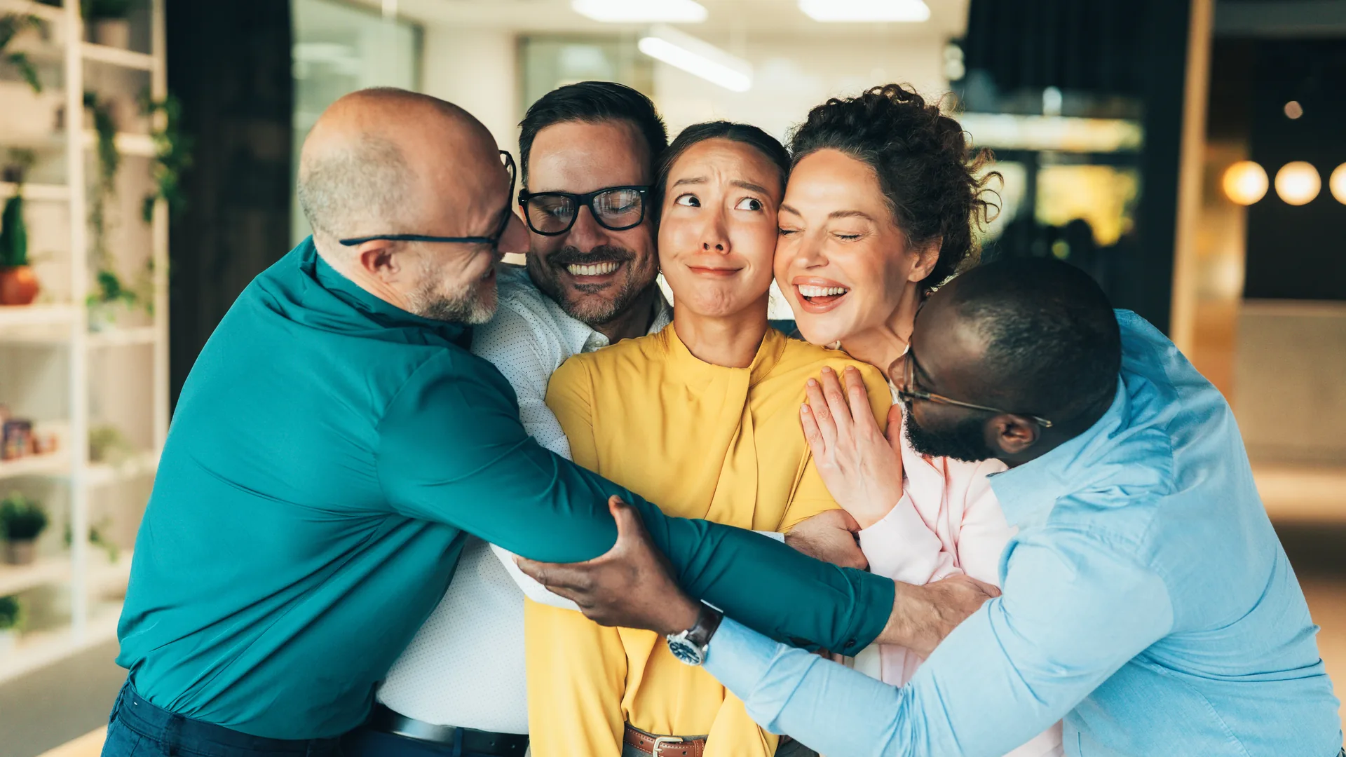 Five colleagues crowding in to hug a woman in a yellow blouse, who pulls a bashful face